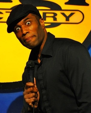 Black comedian Chris James to book for party in a black hat and black jacket making a goofy face holding a microphone in front of a yellow sign.