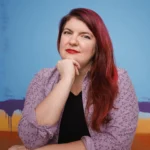 Diversity speaker for hire Madelein Smith with red hair swept to the side, hand on her chin with red lipstick on and mauve button down shirt with a black shirt underneath.