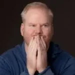 Clean comedian Jim Gaffigan with blue shirt and hands covering his mouth.