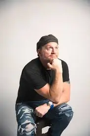 Christian comedian for hire Tim Hawkins with hand on his chin and black shirt and hat on.